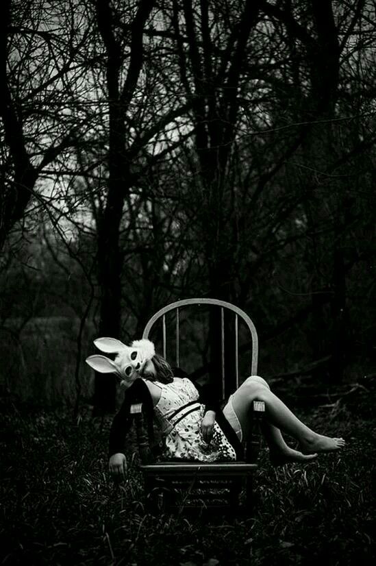 a woman wearing a white bunny mask is sitting on a wooden chair in a dark forest. she is wearing a white dress and has her legs extended. the image is black and white and has a mysterious and spooky atmosphere.