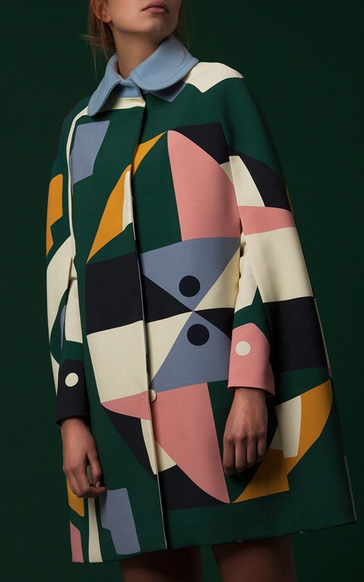 closeup view of a woman wearing a stylish coat. the coat is a kneelength cape with a geometric print in various colors, including green, pink, blue, yellow, white and black. the geometric patterns create an abstract design. the woman is wearing a light blue collared shirt underneath, and the coat has a button closure. the background is a dark green, providing a contrast to the vibrant coat. the image has a fashionoriented style.
