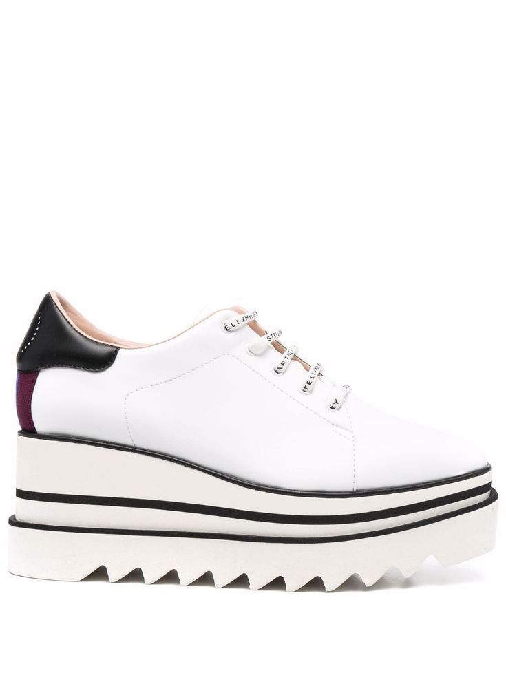 a closeup studio shot of a pair of white leather stella mccartney wedge sneakers. the sneakers have a platform sole with a scalloped edge, black detailing at the heel and a white lace with stella mccartney branding. the shoes are presented on a white background.
