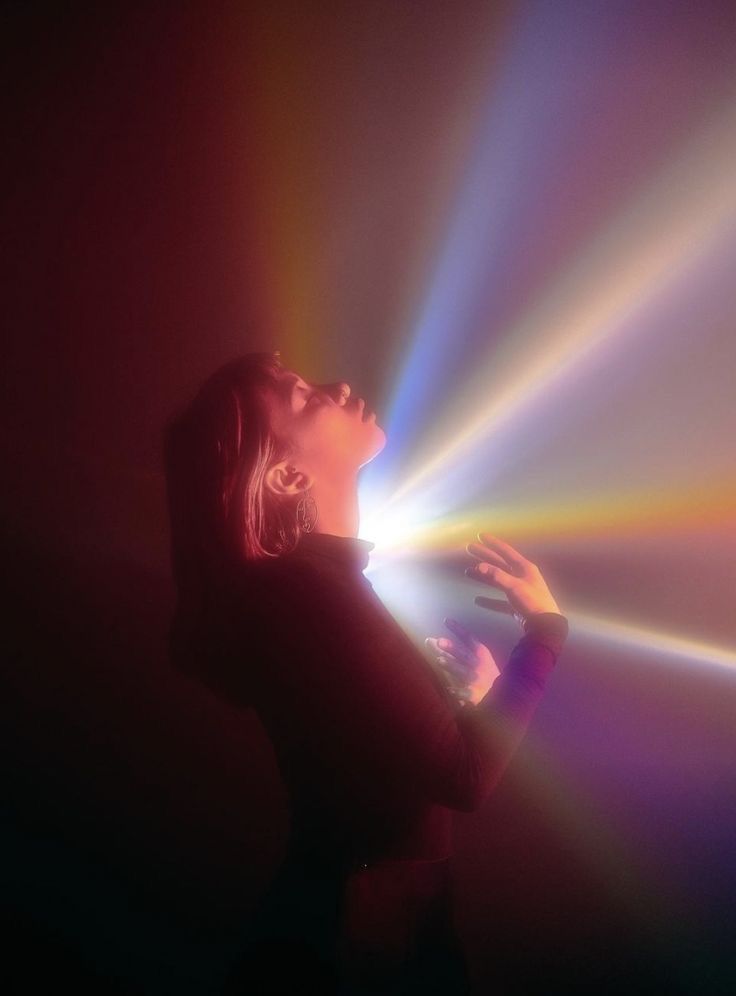 a woman stands with her eyes closed, backlit by a beam of colorful, prismatic light that creates a rainbow effect. her face is upturned and relaxed, as if she's basking in the glow of the light. the image is artistic, ethereal, and dreamy, creating a sense of wonder and mystery.