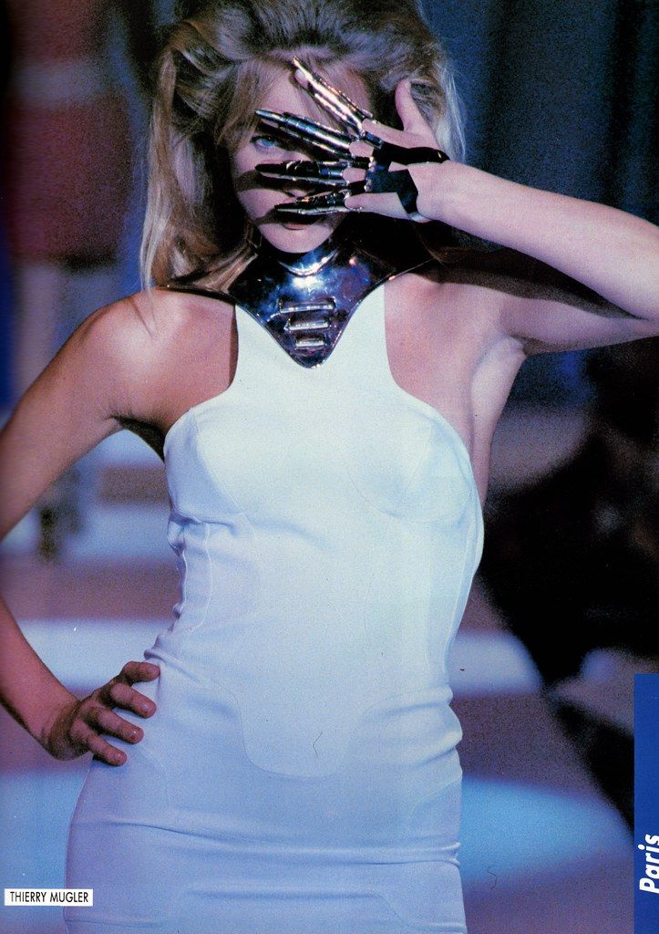 a model in a thierry mugler fashion show in paris is standing on the runway wearing a white dress with a metal collar. she has her blonde hair pulled back and is covering part of her face with her hand, which has metal claws attached to her fingers. the background is blurred with other models and audience members.