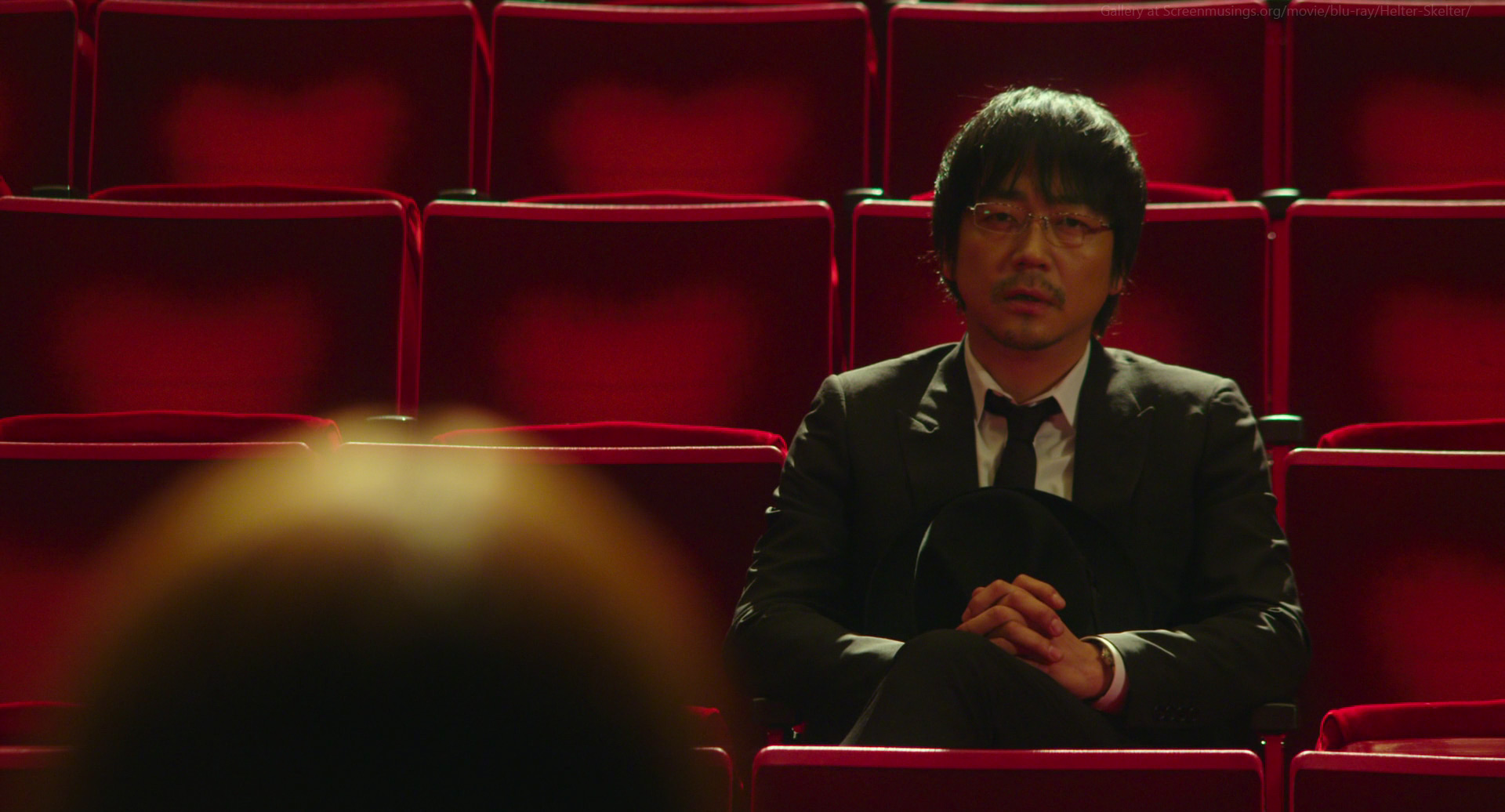 a man in a black suit and glasses sits in a red theater seat. he has a serious expression on his face and is looking down, appearing to be introspective or contemplative. the red seats around him create a stark and dramatic contrast with his attire, emphasizing his somber mood.