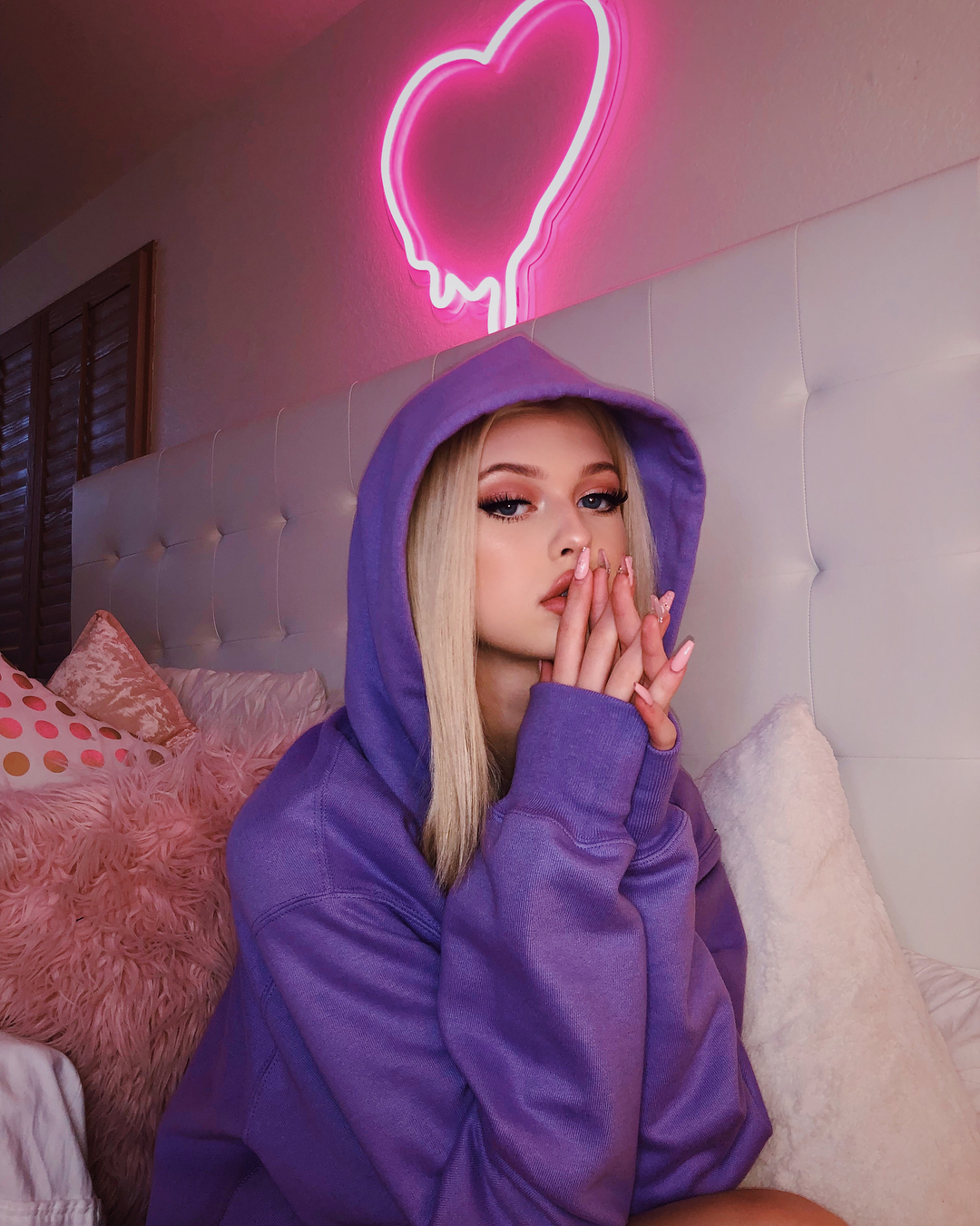 a young woman with blonde hair wearing a purple hoodie is sitting on a bed in front of a white headboard with a pink neon heart sign on the wall above her head. she has her hands clasped together near her face and is looking at the camera. there are decorative pillows behind her.