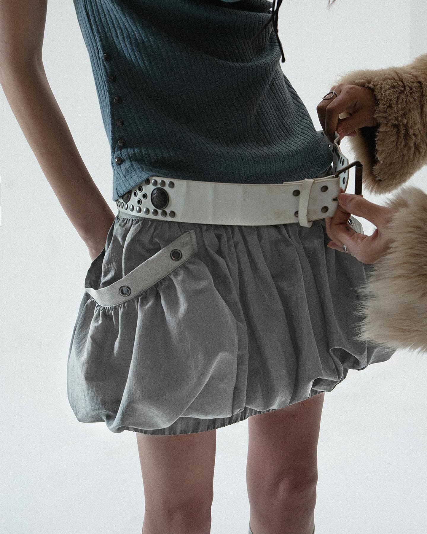 loseup view of a woman modeling a stylish outfit. she's wearing a teal ribbed top and a light grey bubble skirt. a wide white studded belt cinches her waist. another person's hand is partially visible, helping to adjust the outfit. the overall style is modern and slightly edgy. the background is plain white, emphasizing the clothing.
