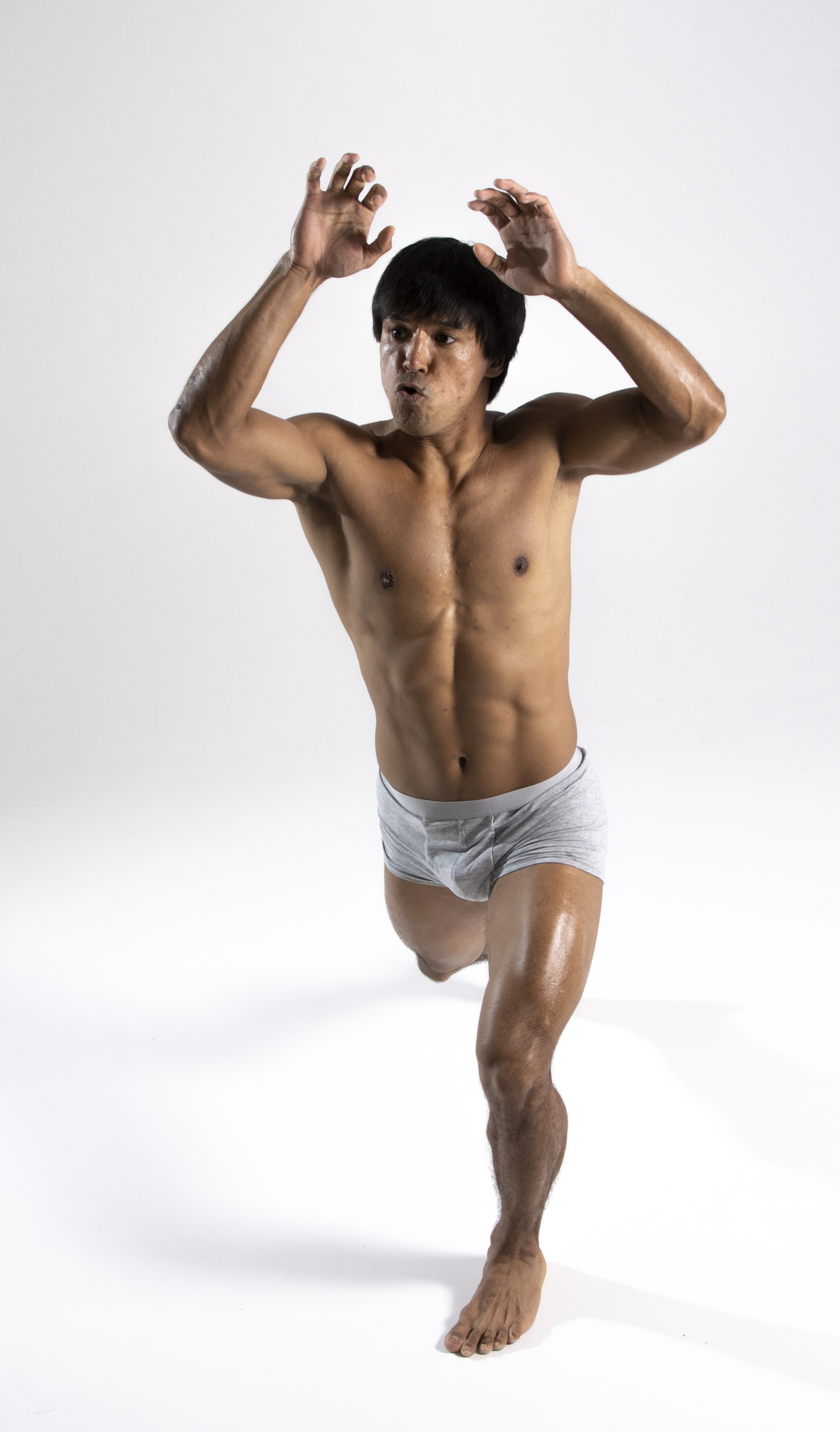 a shirtless asian male is standing in a studio with a white background. he is wearing grey underwear. his arms are raised with his hands up. he is muscular and fit.
