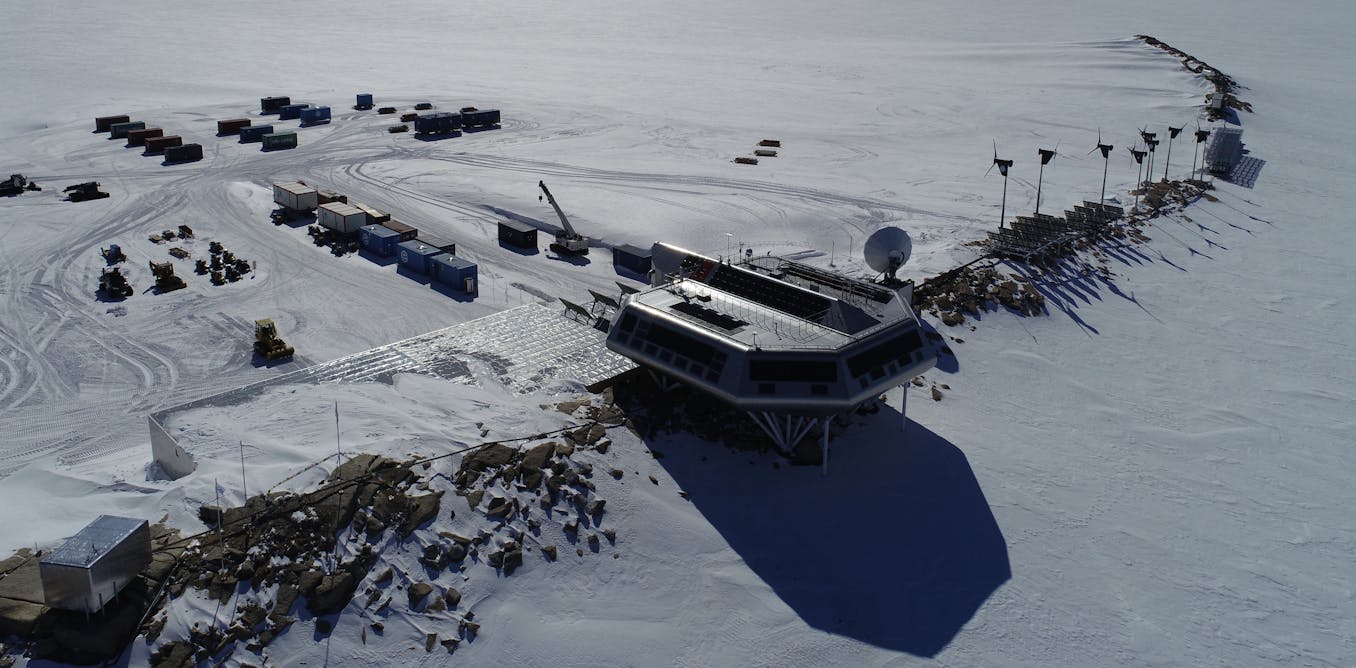 an aerial view of a research station in antarctica. the station is located on a snowcovered landscape and is surrounded by containers, vehicles, and wind turbines. there is a satellite dish on the roof of the station. the sun is shining and the sky is blue. the photo was taken from a helicopter or drone.