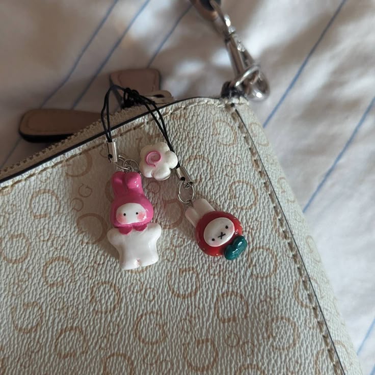 a closeup shows a lightcolored purse with two cartoon character charms hanging from it. one charm depicts the character my melody, white with a pink hood, while the other appears to be a red rabbit with a bluegreen detail. a white flower charm is also visible. the purse has a light pattern and a brown handle attachment, with a metal clasp and chain extending from the top. the background consists of light blue and white striped fabric.