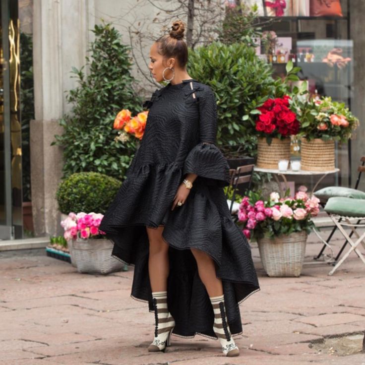 a woman in a black, highlow dress with ruffled details is standing outdoors on a brick path. she has her hair in a bun and is wearing hoop earrings, jewelry, striped socks, and heeled boots. there are flower arrangements behind her.