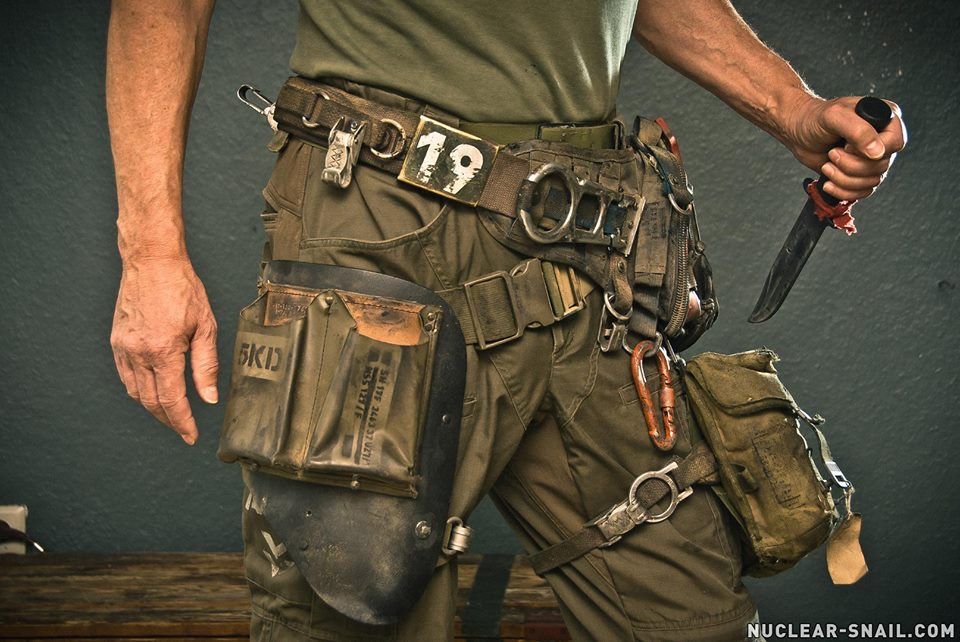 a closeup shot of a man's hand, holding a knife. he is wearing a tool belt with a variety of tools attached to it, and he is standing in front of a green background. he looks like a construction worker or someone who works outdoors. the picture is a closeup and focuses on the tools and the man's hand. the background is blurred.