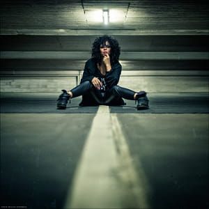 a young woman sits in the middle of an urban street. she is wearing a dark jacket and jeans with boots and has her hair pulled back and is looking away.