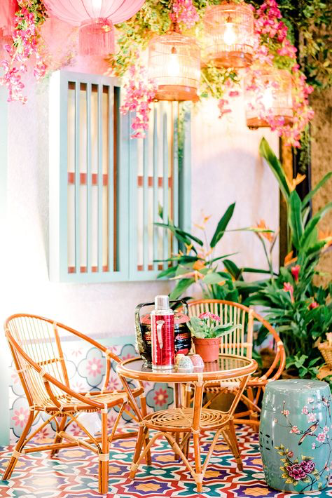 a charming outdoor patio scene featuring two rattan chairs surrounding a small round glasstopped table. the setting is lush with tropical plants and pink flowers, enhanced by hanging chinese lanterns adding a whimsical touch. the floor is covered in vibrant patterned tiles, and the wall is a soft teal. a decorative ceramic stool with bird and floral designs adds to the aesthetic. the overall atmosphere is calm, inviting and aesthetically pleasing, suggesting a cafe or restaurant setting.