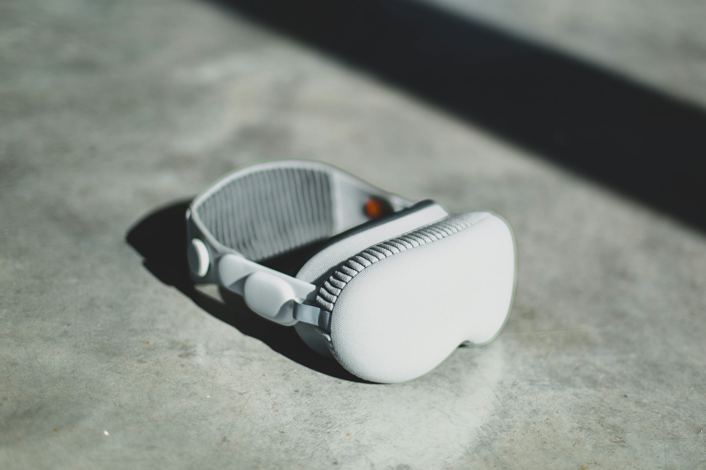 a pair of gray apple airpods max headphones are lying on a gray concrete surface. the headphones are positioned at an angle, with the headband and earcups clearly visible. a shadow is cast behind the headphones, and there is a streak of light in the background. the concrete surface is plain and slightly textured.