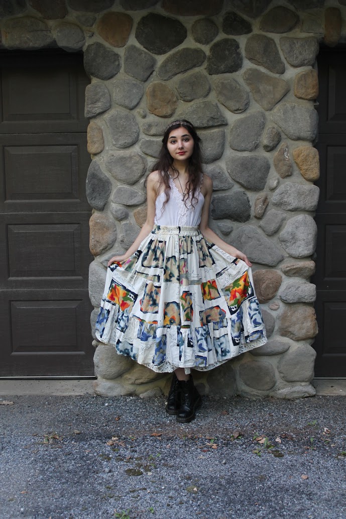 a young woman is standing in front of a rock wall wearing a white top and a long, colorful patchwork skirt. she is wearing black boots and a headband. the scene is outdoors.