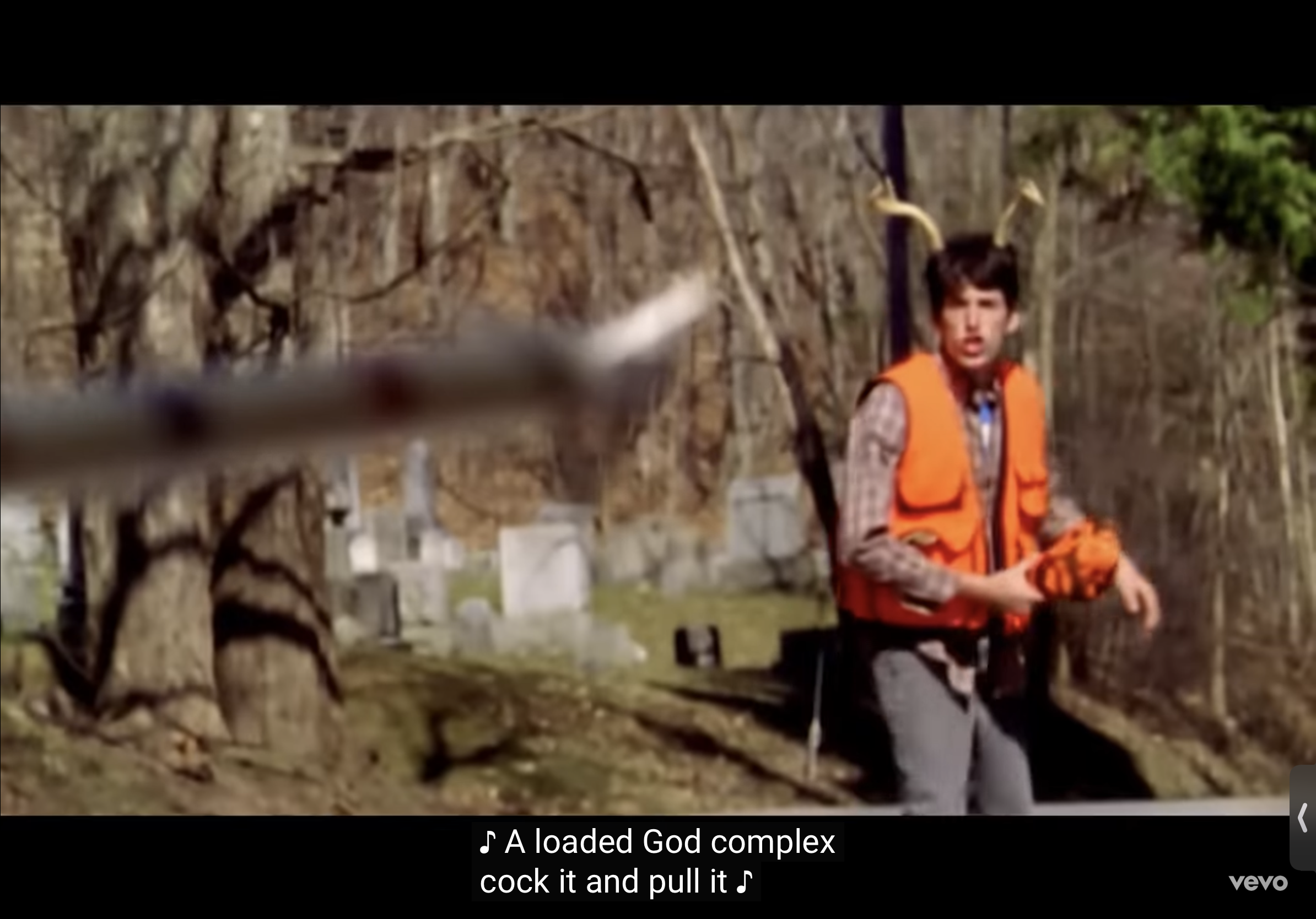 a man wearing an orange vest stands in a forest. he has a gun in his hand and is looking at something in the distance. there are trees and leaves in the background and the man is standing near a cemetery.