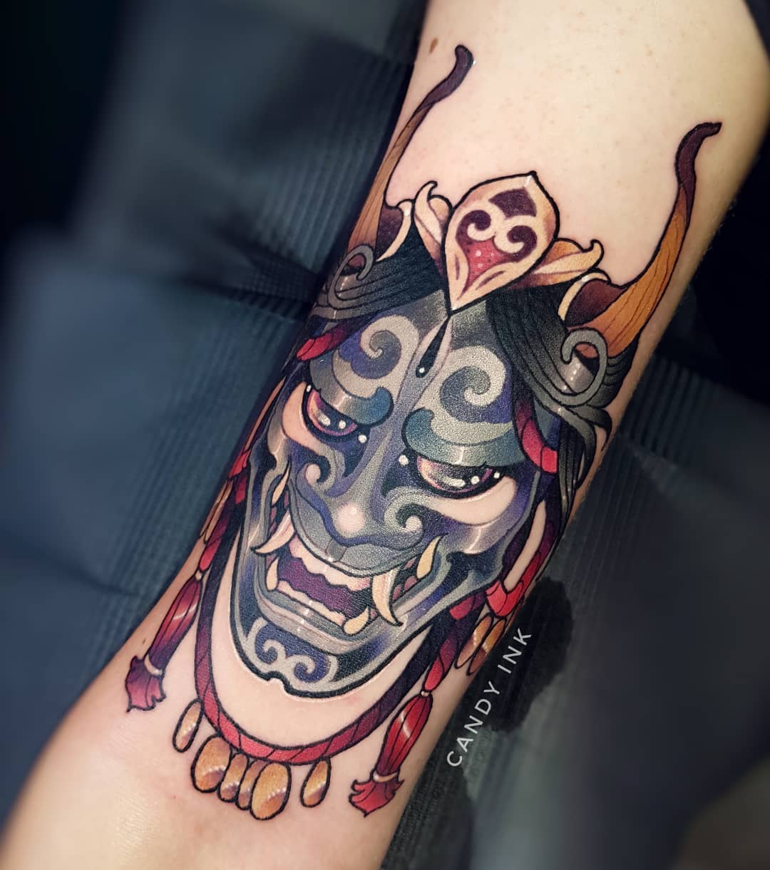 a closeup photograph showcasing a vibrant neotraditional tattoo of an oni mask on the back of someone's forearm. the tattoo is rendered in a bold and graphic style, with thick black outlines and a rich color palette. the oni mask is depicted with its characteristic fierce expression, with sharp horns, bulging eyes, and a gaping mouth. the tattoo also features decorative elements such as swirling patterns and a string of beads, adding to its visual appeal.