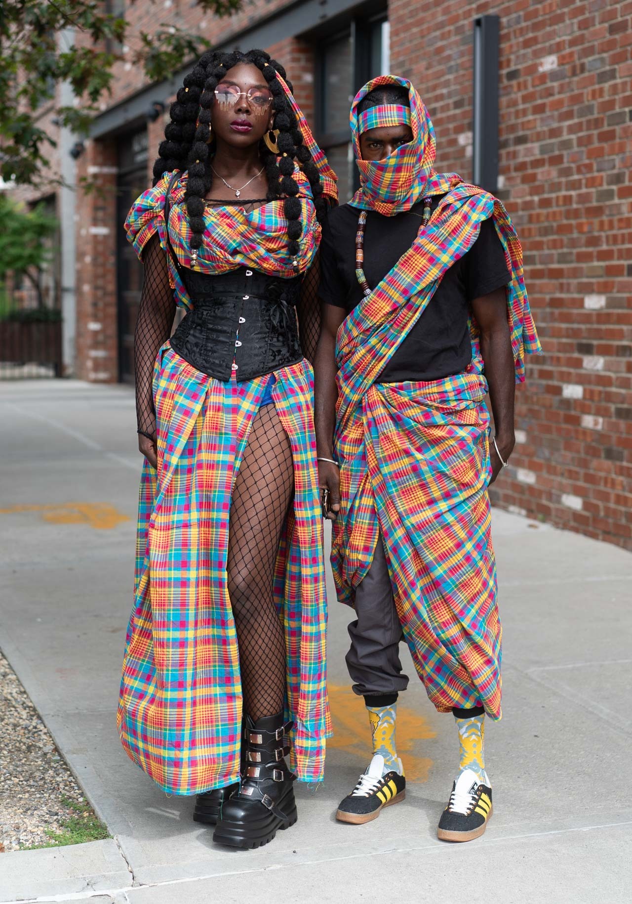 a fashionable couple walks down a city sidewalk. the woman wears a long, plaid dress, a black corset, and chunky black boots. she has long black hair with large curls. she wears sunglasses. the man wears a similar plaid kilt, a black top, and sneakers. he wears a patterned scarf over his face. they both appear to be african.  the couple is confident and stylish.  they are walking toward the camera. the background is a brick wall. there are some green plants in the bottom left corner.