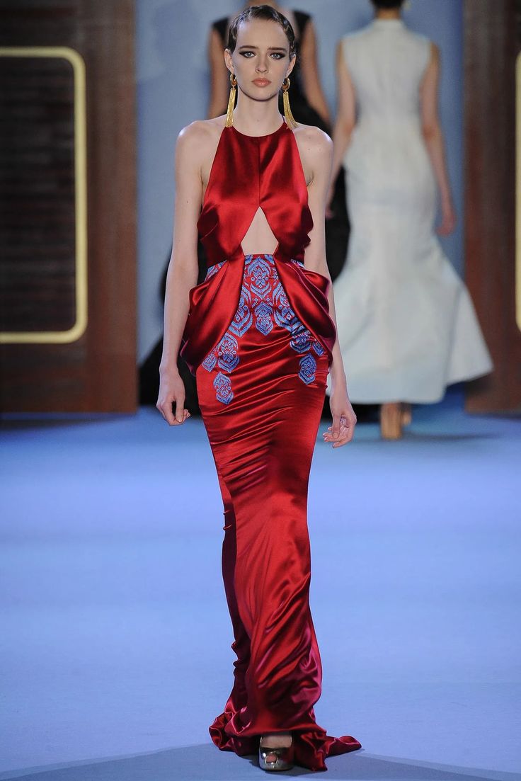 a woman in a red gown with blue embroidery is walking the runway. she has her hair pulled back and is wearing gold earrings. she is looking straight ahead, and her expression is serious.