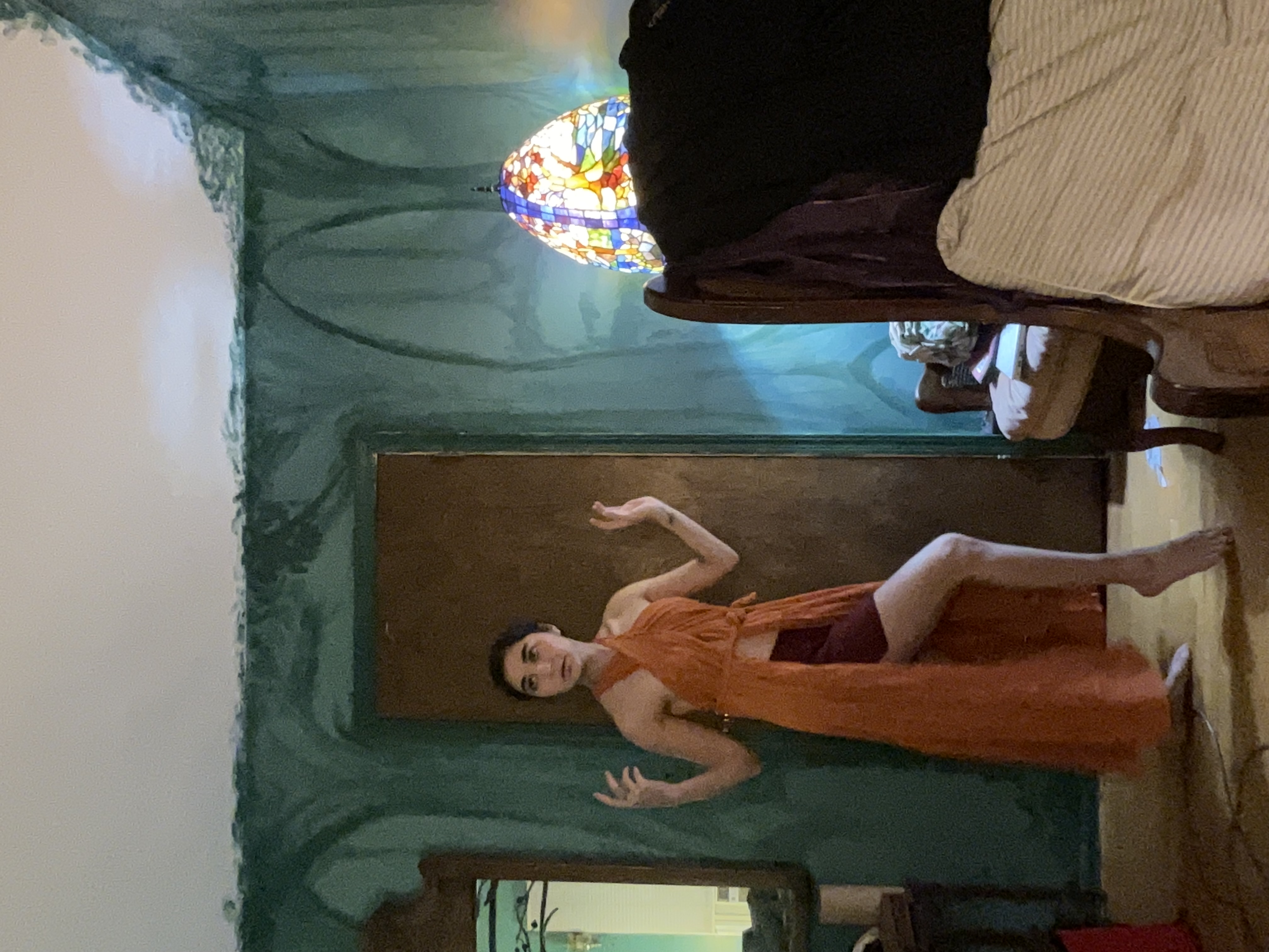 a fulllength shot of a woman in a long, orange dress posing in a room with teal painted walls. the walls have a painterly, almost abstract design. she is standing with her arms slightly raised, one leg slightly bent.  an armchair and the edge of a bed are visible in the background, along with a stainedglass lamp. the overall aesthetic is vintage or retro, with an artistic feel. the lighting is soft and somewhat subdued.