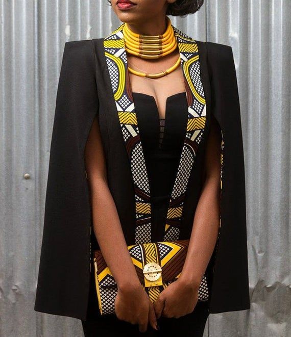 the image showcases a fashionable woman wearing a stylish black cape blazer with a contrasting lining featuring a vibrant yellow and brown african print (likely ankara or kitenge). the cape is sleeveless and falls elegantly to midthigh.  she is accessorized with a large, chunky gold and ambertoned necklace that complements the colors of her outfit and a matching clutch bag made from the same patterned fabric as the blazer's lining. the woman's pose and confident expression contribute to the overall sophisticated and elegant feel of the image. the background is a neutral corrugated metal wall, allowing the outfit to be the focus.