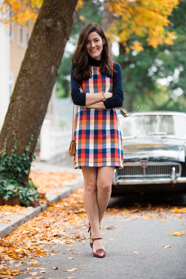 a smiling woman in a colorful plaid dress stands on a street covered with autumn leaves.  she's wearing a dark turtleneck, brown shoes, and a small brown handbag.  a vintage car is parked behind her. the overall scene evokes a feeling of autumnal charm and retro style.