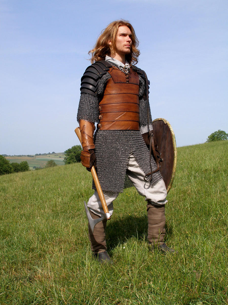 a man dressed in medievalstyle armor, including chainmail and a leather tunic, stands in a grassy field. he carries an axe and a shield. the setting appears to be outdoors on a sunny day. the man's long hair and the overall style suggest a fantasy or historical reenactment setting. he's looking away from the camera.