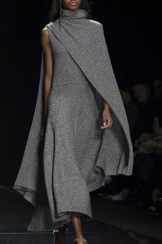 a fashion model walks down the runway in a long, flowing silver dress. the dress is made of a soft, shimmery fabric and has a cape that drapes over the model's shoulders. the model is wearing high heels and her hair is pulled back. the background is dark, which makes the model's dress stand out even more.
