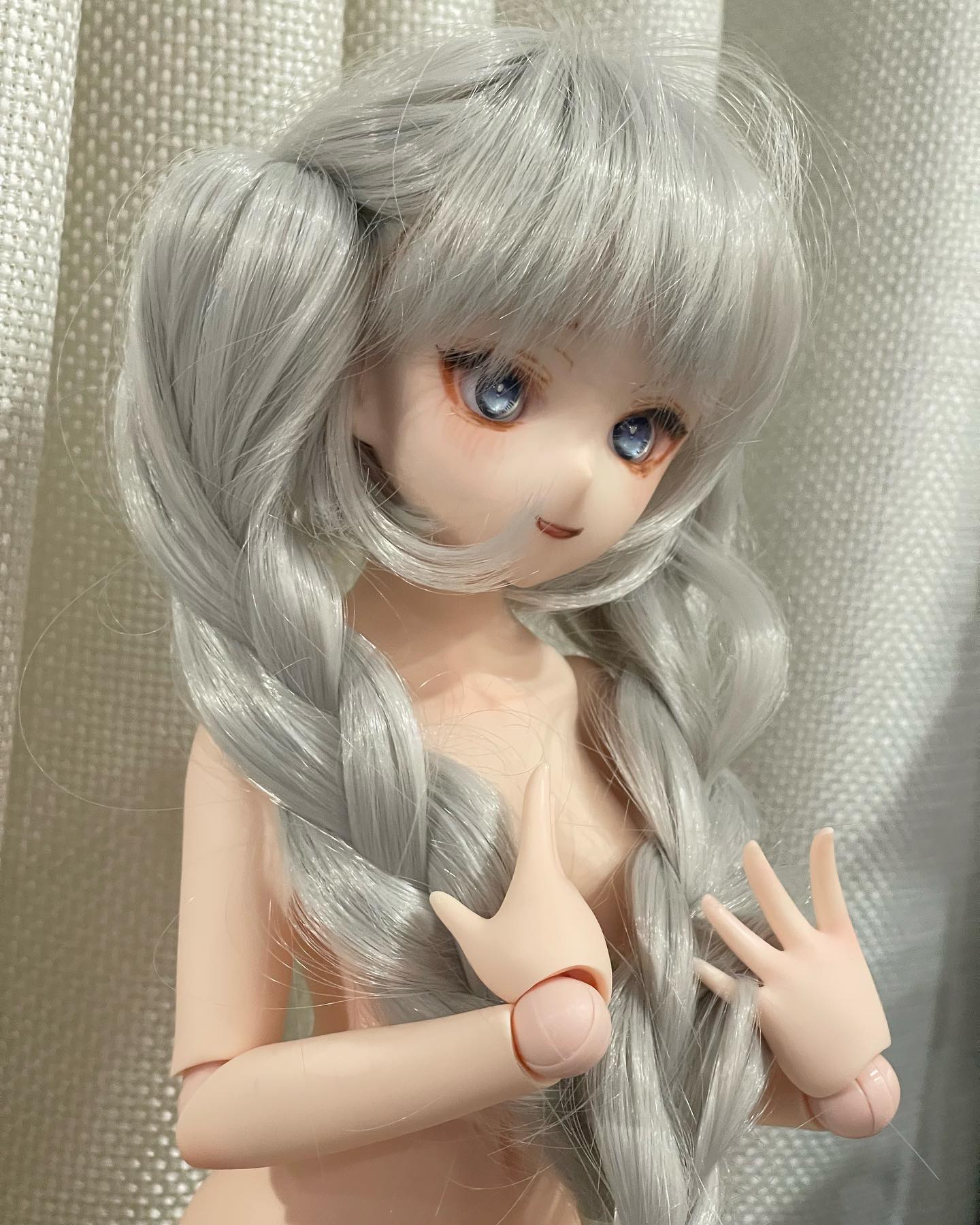 a closeup image of a balljointed doll with grey braided hair, styled in two pigtails. the doll has blue eyes with orange eyeshadow and a soft pink lip. the doll is nude and positioned in front of a white textured curtain.