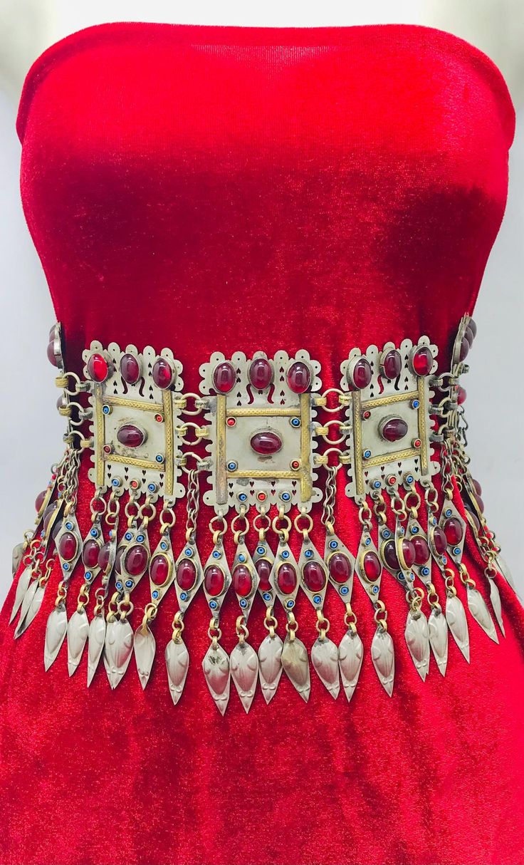 a closeup shot of a decorative, elaborate necklace draped over a red velvet display. the necklace features a silver and gold metallic design with several red gemstones and pendants. the piece has a vintage, tribal aesthetic, likely from afghan or similar cultural influences, given the design and style of the ornamentation.