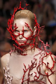 a woman with a corallike headpiece and red lipstick is wearing a coralinspired dress, showcasing a futuristic and avant garde fashion design. the headpiece is made of red coral and covers her head and face, leaving only her eyes and lips visible. the corallike dress follows the curves of her body and creates a unique silhouette. the overall design is creative and artistic, blending nature with modern fashion.