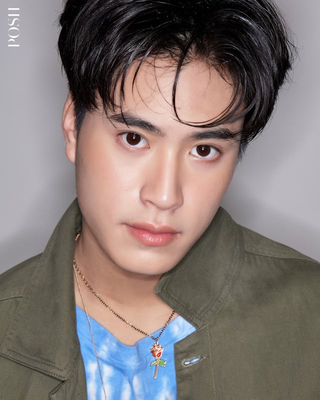 a closeup portrait of a young asian man with short black hair. he is wearing a green overshirt, a tiedye blue and white shirt underneath, and a gold chain with a red rose pendant. he has brown eyes and a neutral expression. the background is a light gray. the lighting is soft and flattering.