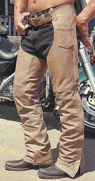 the image shows a man wearing tan leather chaps over black denim jeans. he is standing next to a green and chrome cruiserstyle motorcycle.  the chaps are fastened with buttons at the bottom of each leg. the man is wearing black motorcycle boots and has a belt around his waist. the overall setting suggests a biker or motorcyclist lifestyle.