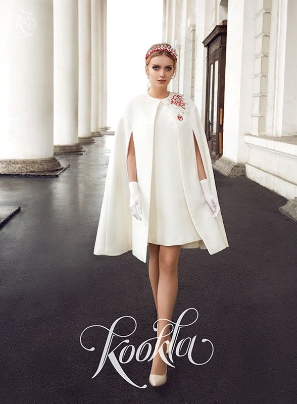 the image showcases a fashion model wearing a stylish, elegant white ensemble.  the outfit consists of a short, aline white dress adorned with subtle red embroidery on the bodice, paired with a flowing white cape that extends below the dress's hemline. the model wears white gloves and highheeled shoes.  she's accessorized with a crystal headband. the background features classical architecture with white columns, creating a sophisticated and timeless setting. the "kookla" brand logo is subtly overlaid on the image. the overall aesthetic suggests a bridal or special occasion look, possibly wedding attire.