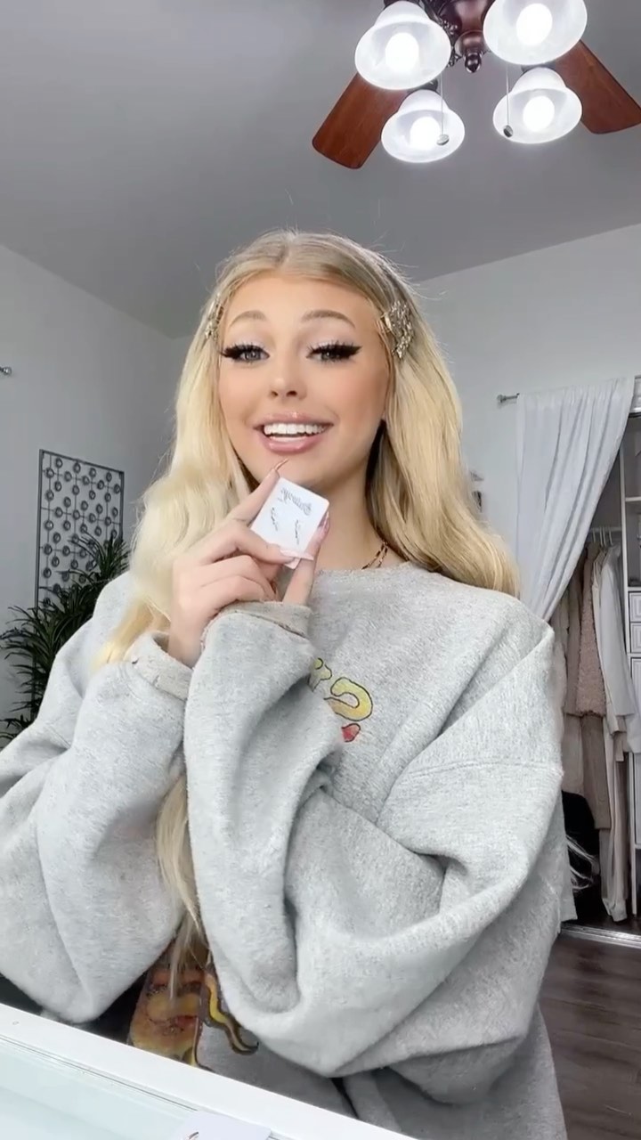 a smiling woman with long blonde hair and hair clips is holding a small white makeup compact. she is wearing a gray sweater. in the background is a ceiling fan, a houseplant, and a closet.