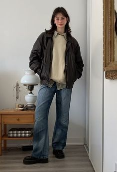 a young woman stands casually in a room, posed with her hand in her pocket. she is wearing a black leather jacket over a light brown sweater, dark wash denim, and black boots. the woman is standing against a white wall with a white floor, next to a wooden nightstand with a lamp. a wooden mirror is hanging on the wall in the background.