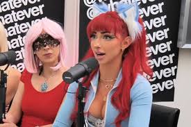 the image shows a closeup of three women seated in what appears to be a podcast studio setting.  the central figure has bright red hair styled with animestyle cat ears and a matching headband. she is wearing a light blue zipup jacket, revealing a lowcut top.  she is positioned between two other women. the woman to her left has bright pink hair and is wearing a masqueradestyle mask and a red dress.  the third woman (partially visible on the left) has light blonde hair. two microphones are visible, suggesting the scene is a recording of some kind. the background features a wall with the word "whatever" repeated multiple times.  the overall atmosphere suggests an interview or podcast recording featuring cosplayers or women in costume.