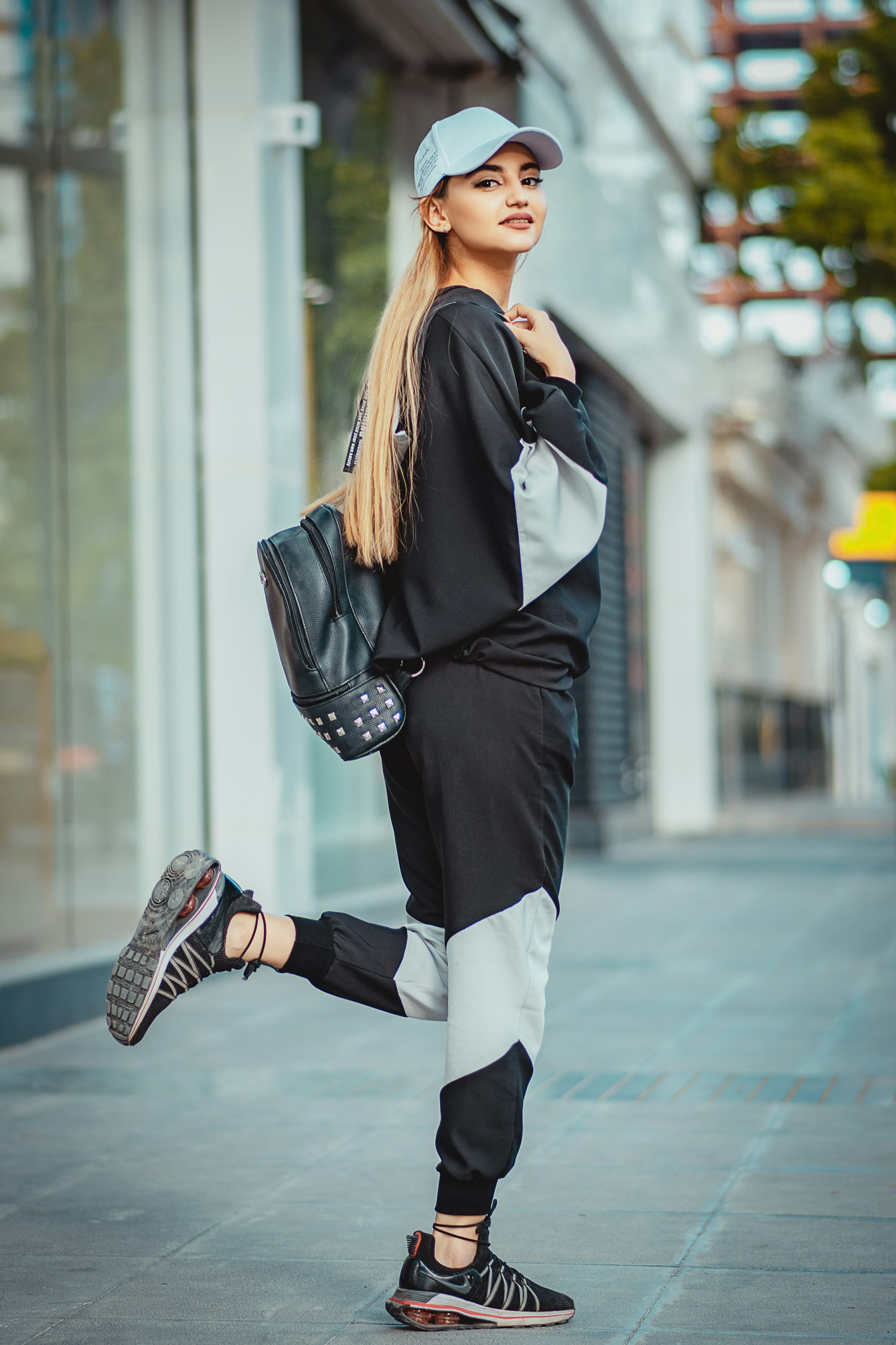 a young woman in stylish streetwear poses on a city sidewalk. she's wearing a black and white jogger set, a light blue baseball cap, and black sneakers.  she carries a black backpack and has long blonde hair. the overall style is casual and trendy, perfect for active wear or everyday urban style. the photo has a bright and cheerful feel, suggesting a positive and confident attitude.