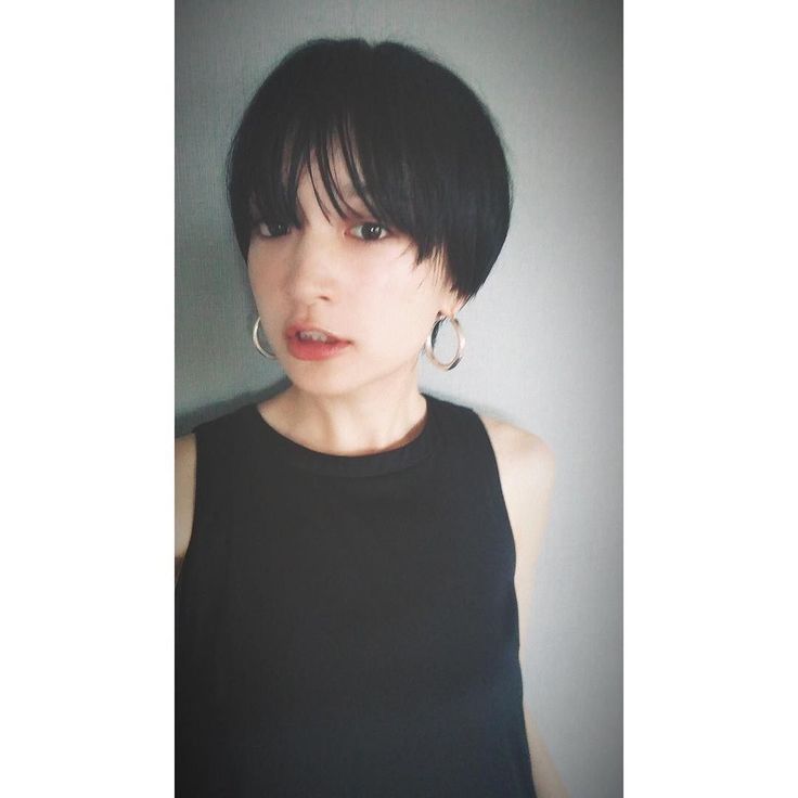 a young woman with short black hair, styled in a bob with bangs, is looking at the camera. she is wearing a black sleeveless top and silver hoop earrings. the background is a plain gray wall.