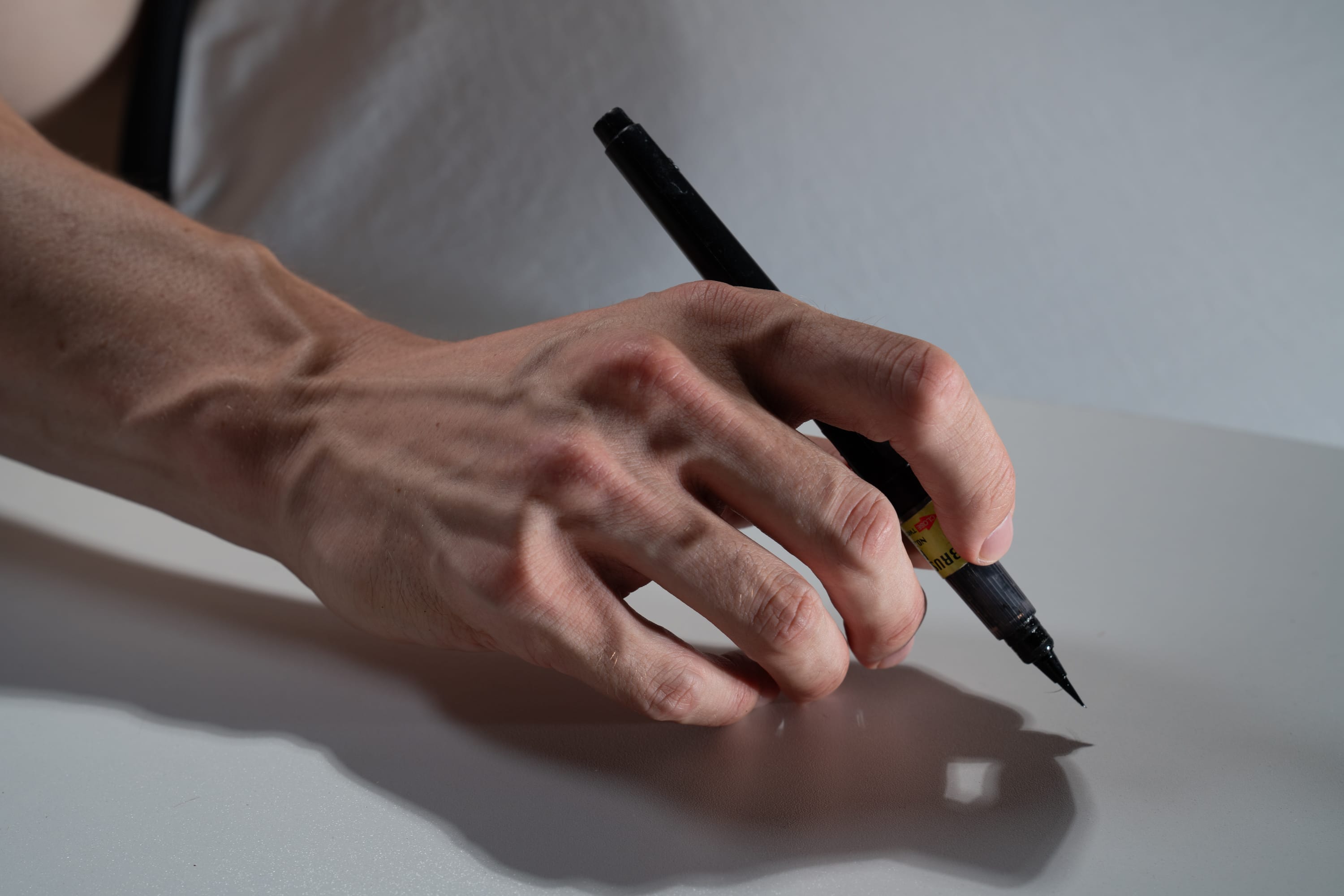 a closeup shot of a man's hand holding a black brush pen poised above a white surface. the image suggests an action about to happen, like the beginning of a writing or drawing session. the studio lighting emphasizes the hand's details and the pen's shadow on the surface.