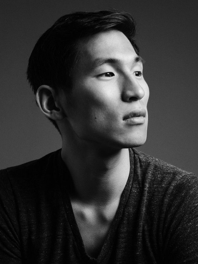 a black and white portrait of a young asian man looking up with a serious expression. he is wearing a dark vneck tshirt and has short black hair. the photo was taken in a studio setting.