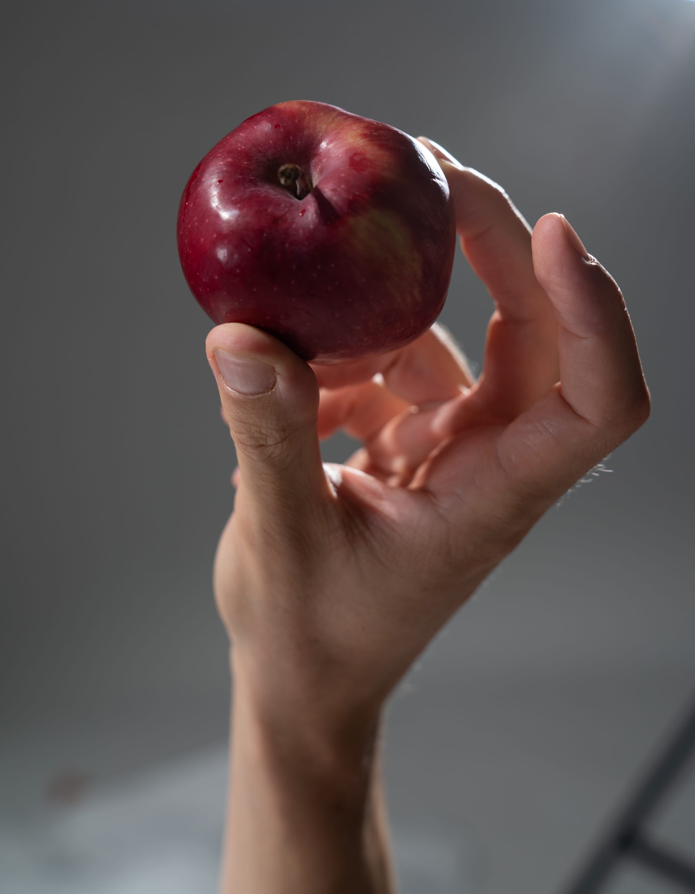 a hand holding a red apple with a stem in front of a gray background.