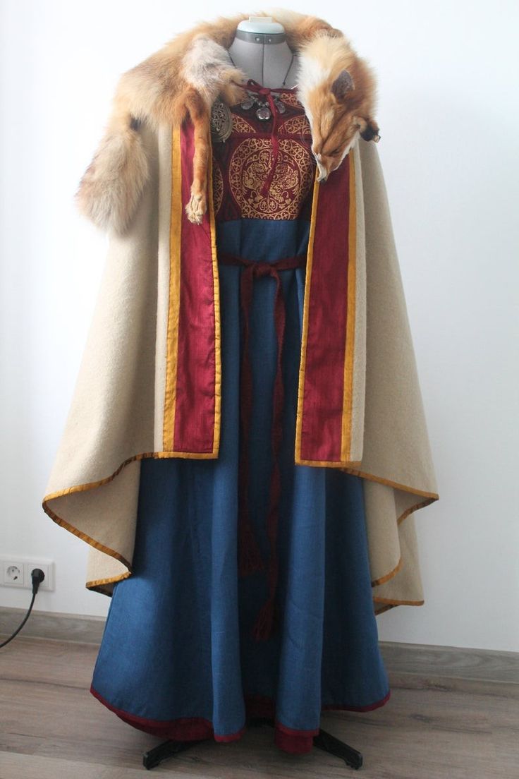 a woman is wearing a medieval viking dress, complete with a cloak, fur, and jewelry.  the dress is a dark blue color, and the cloak is a lighter beige color with red and gold trim.  the woman is also wearing a fur collar and necklace. the costume is historically accurate.