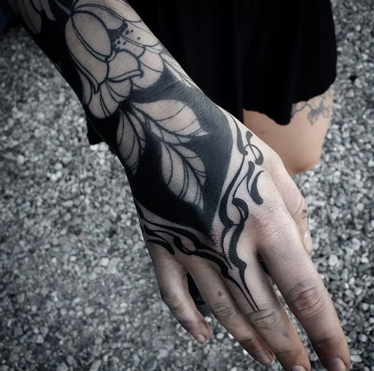 the image shows a closeup of a hand and forearm featuring a blackwork tattoo with a floral design. the tattoo extends from the fingers to the forearm, encompassing a flower with leaves and intricate patterns. the majority of the tattoo is solid black ink with line work on the flower petals and leaves.