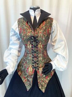 a closeup view of a mannequin dressed in a historicalinspired outfit. the focus is on the ornate green, gold, and black tapestry vest with a laceup front. the vest is paired with a crisp white blouse with puffed sleeves and a black collar, hinting at a victorian or renaissance aesthetic. the bottom half of the outfit is not visible.