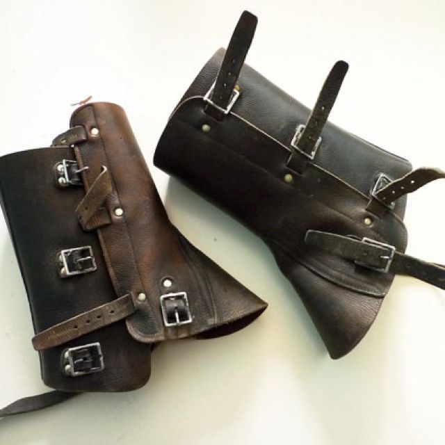 two brown leather boot protectors or armor pieces for the lower legs. both are laced with thick straps and buckles and are made of supple thick leather. they are lying on a plain white background.