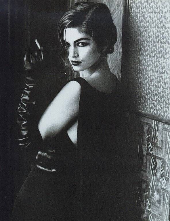 a black and white portrait of a woman in a black dress and gloves. she is standing in a dark room and looking directly at the camera. her hair is styled in a glamorous way and she is wearing makeup. the woman's expression is serious and her stare is seductive. the image is dark and mysterious, with the woman's face and body illuminated in a soft light. the overall effect is one of elegance and sophistication.