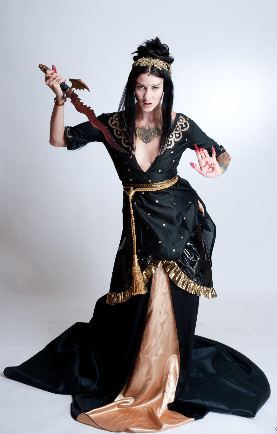 a fullbody studio shot of a woman in a dramatic black and gold gown. she is holding a dagger and her pose suggests power and mystery. her makeup is striking, and she has henna or similar markings on her hands. the background is plain and light, allowing the woman and her attire to be the focus of the image.