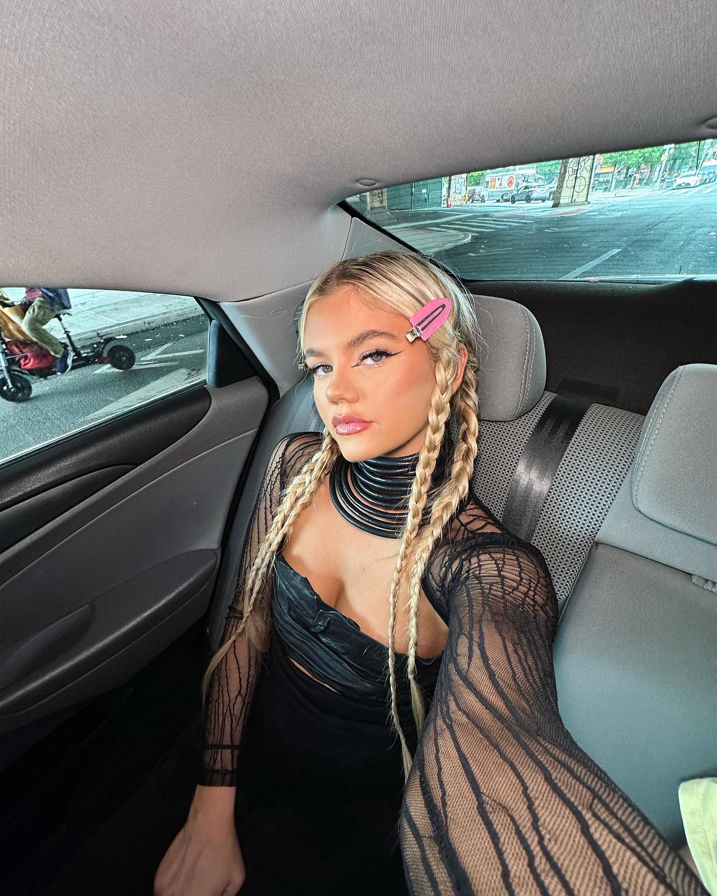 a woman with blonde hair styled in braids and a pink hair clip sits in the passenger seat of a car. she is wearing a black dress and a choker. the car is moving along a street, visible through the window. the woman is taking a selfie.