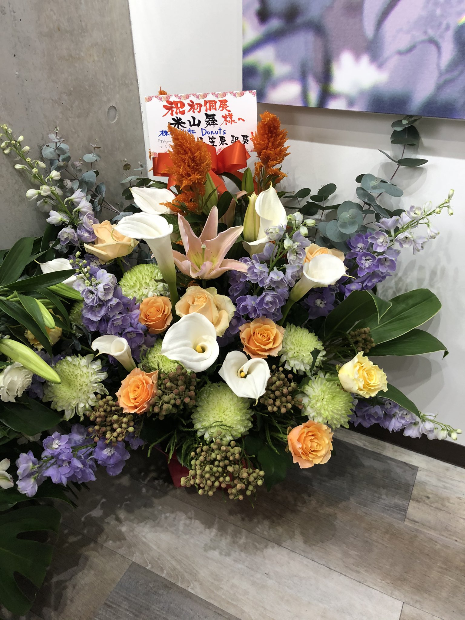a colorful floral arrangement sits on the floor in front of a wall, possibly celebrating the grand opening of a business.  the arrangement features lilies, roses, and other colorful flowers in shades of orange, yellow, purple, and white. the sign on the arrangement contains japanese characters.