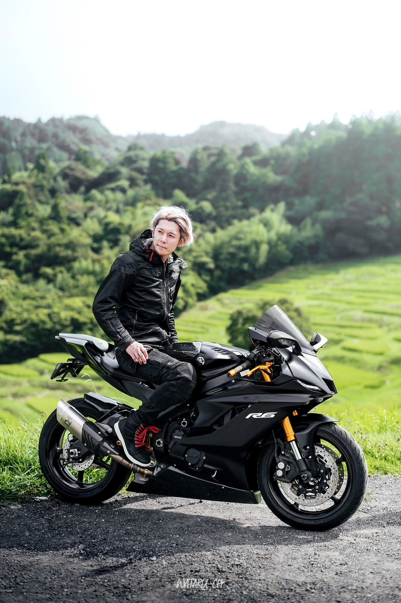 a woman in black leather gear sits on a black yamaha r6 sportbike, staring out into a mountainous landscape. she is wearing a black leather jacket, black pants, and black gloves. the bike is parked on a paved road, with green grass and hills in the background. the sun is shining brightly, and the image is taken from a low angle. the woman appears to be on an adventure, and she looks both powerful and free.