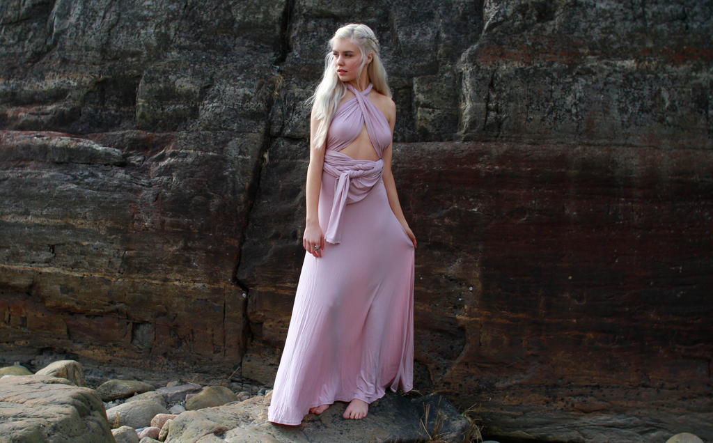 a young blonde woman in a long, flowing, dusty rose maxi dress stands barefoot on a rocky beach.  the dress is a halter style with a wrap design.  she has long blonde hair in a braid. the background is a dark, textured cliff face. the overall feeling of the image is elegant and serene.